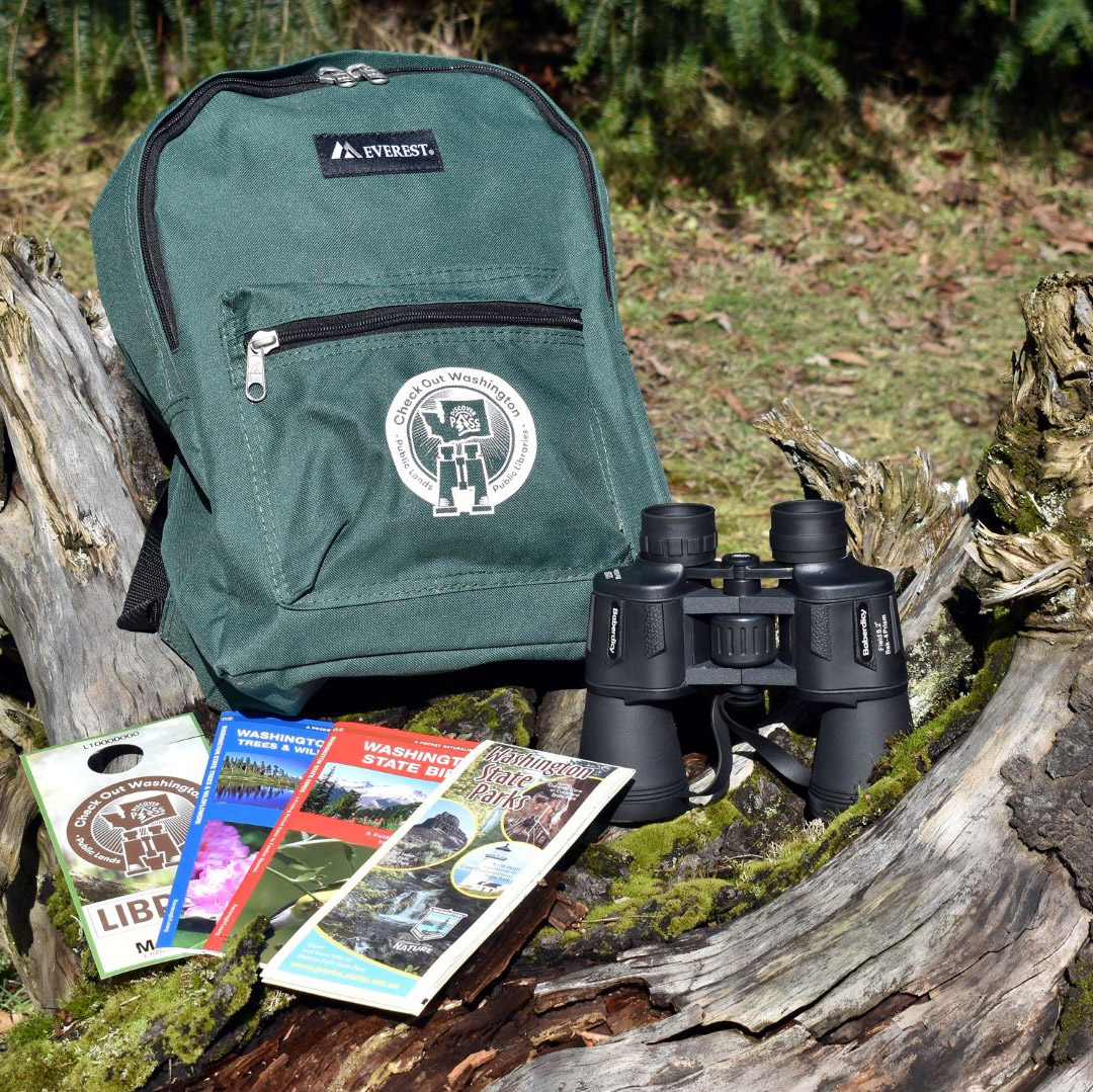 Check Out Washington backpack, binoculars, Washington Birds pamphlet, Washington Trees and Wildflowers pamphlet, a Washington State Parks guide, and Discover Pass displayed on a log